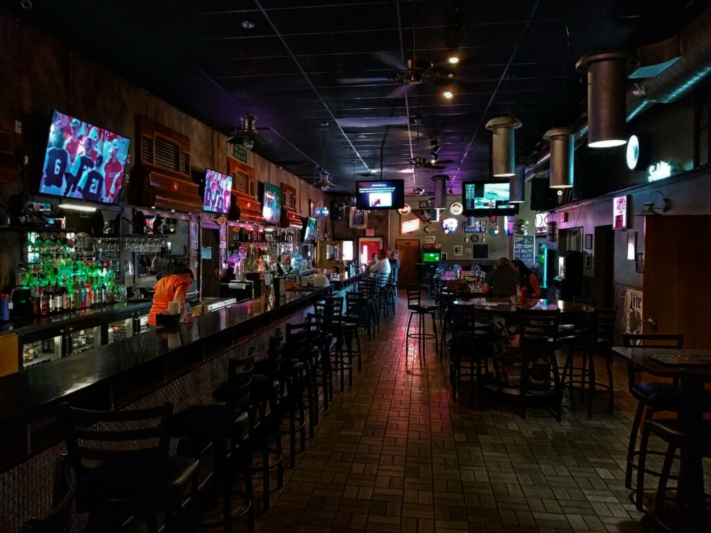 A dimly lit modern bar interior featuring a long counter and multiple screens, creating a cozy urban atmosphere.